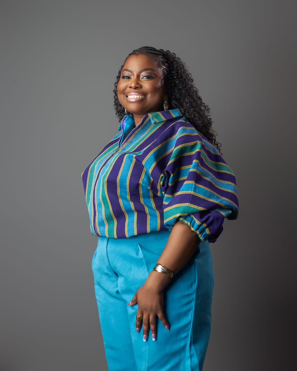 Full-length studio portrait of Joan Adu in a striped heritage-textile blouse and turquoise trousers.
